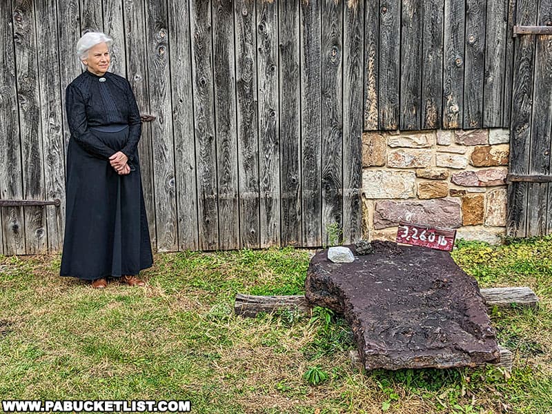 A docent in period costume standing next to what was the last piece of cast iron produced at Curtin Village.