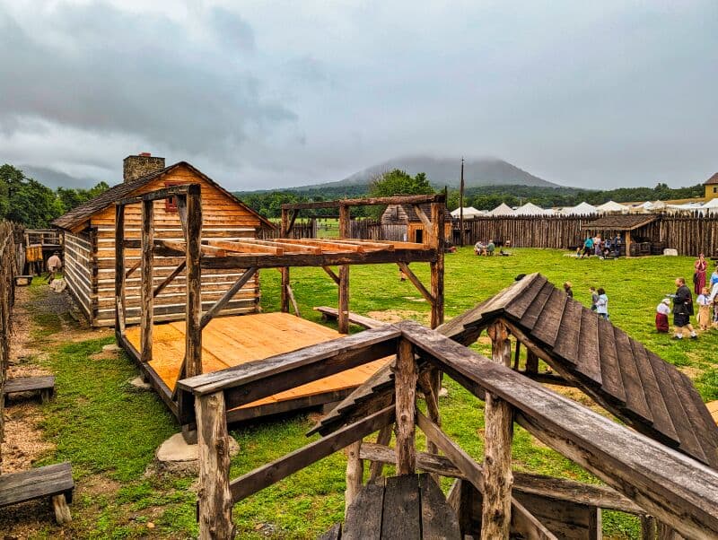 Rustic wooden buildings and elevated platforms inside Fort Loudoun in Franklin County, Pennsylvania. Visitors in period clothing explore the fort, with a misty mountain backdrop and a cloudy sky overhead