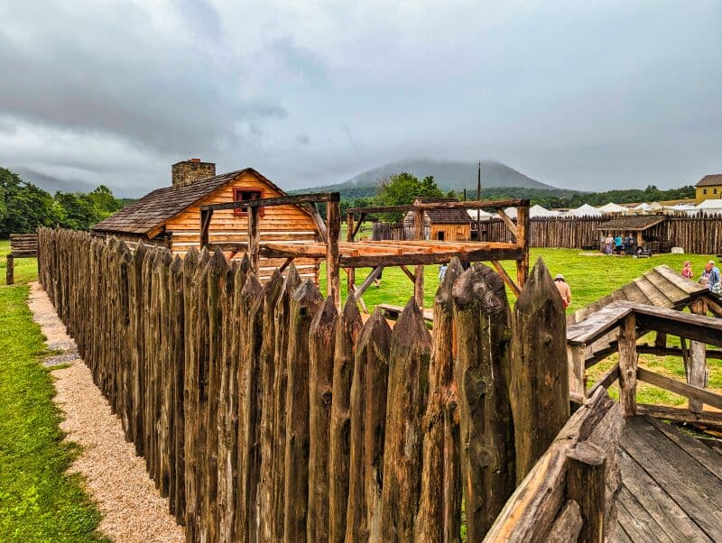 View along a rugged wooden palisade at Fort Loudoun in Franklin County, Pennsylvania. Behind the fence are historical wooden structures, visitors in colonial attire, and a mist-covered mountain in the distance under a cloudy sky.