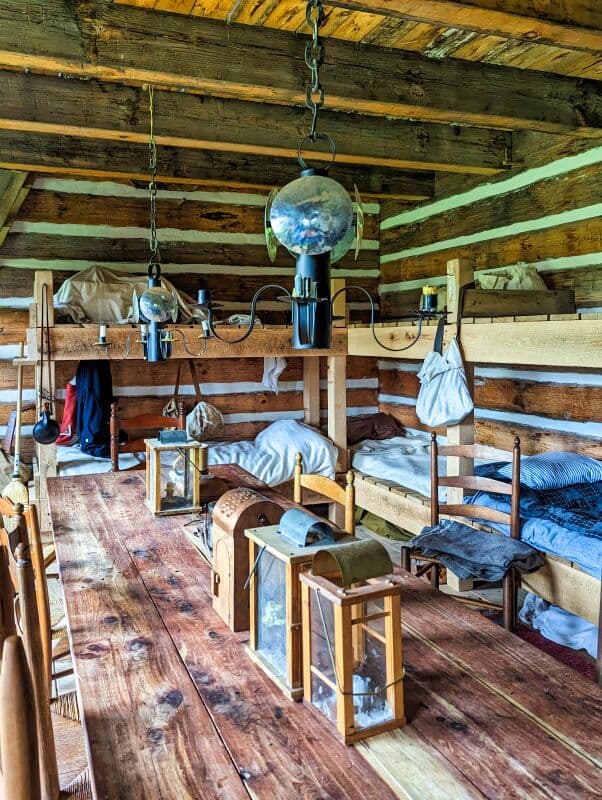 Interior view of a barracks room at Fort Loudoun in Franklin County, Pennsylvania. Features wooden bunk beds with white linens, rustic lanterns placed on a plank floor, and a chandelier-style lamp hanging from the ceiling.