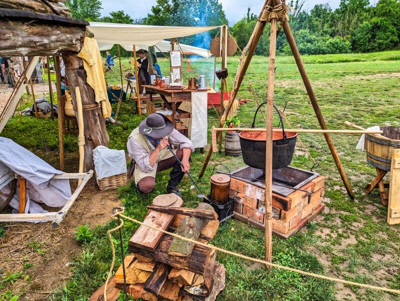 Reenactor in colonial attire tending to a campfire and cooking pot under a canvas tent, surrounded by historical cooking utensils and wooden crates, at Fort Loudoun in Franklin County, Pennsylvania.