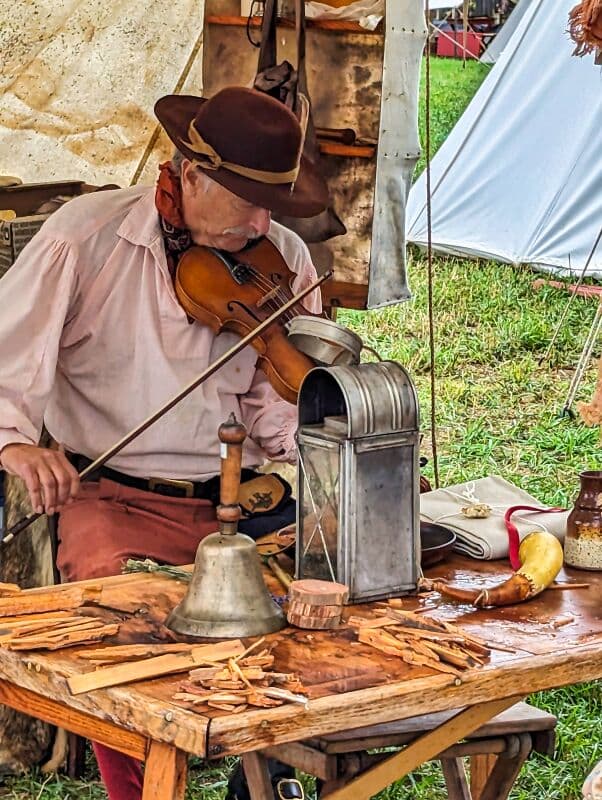 Man in period clothing playing a fiddle while seated at a wooden table displaying various historical artifacts, including a metal container and bell, amidst tents at Fort Loudoun in Franklin County, Pennsylvania