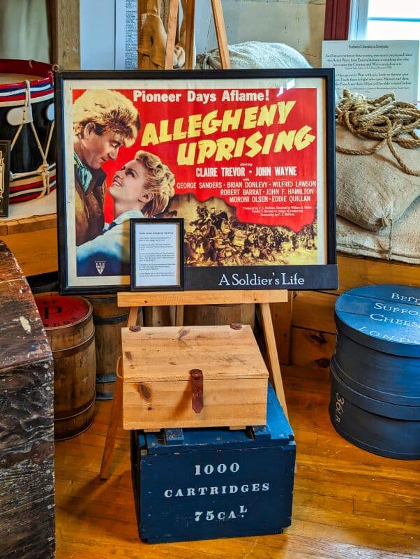 Vintage movie poster titled 'Allegheny Uprising' featuring actors Claire Trevor and John Wayne, displayed alongside a sign reading 'A Soldier's Life', historical artifacts, wooden crates, and barrels in an exhibit at Fort Loudoun in Franklin County, Pennsylvania