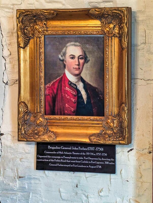Portrait of Brigadier General John Forbes (1707-1759) framed in an ornate gold frame. He is depicted wearing a red military coat with a white cravat. Below the portrait, a plaque provides historical details about his role as the Commander of Mid-Atlantic Theater of the French and Indian War and his contribution to the construction of the Forbes Road. The portrait and plaque are set against a weathered white brick wall at Fort Loudoun in Franklin County, Pennsylvania.