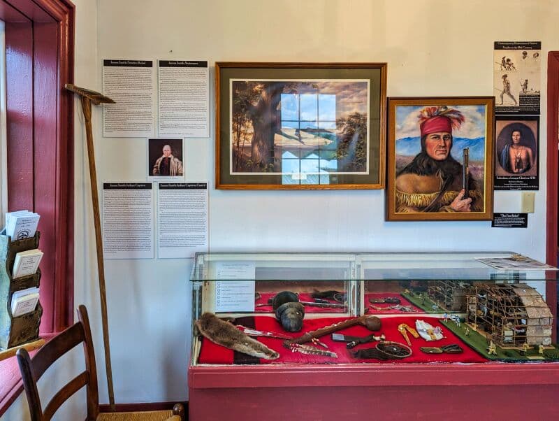 Interior of a museum room at Fort Loudoun in Franklin County, Pennsylvania. The room features a display case filled with historical artifacts such as weapons, tools, and a model of a fort. On the walls, there are framed paintings, one of a Native American man in traditional attire and another of a serene landscape with a flying bird. Multiple informative placards are affixed to the walls, detailing the history and significance of the items and personalities associated with the fort. An old-fashioned wooden chair and a wooden pole lean against the left wall.