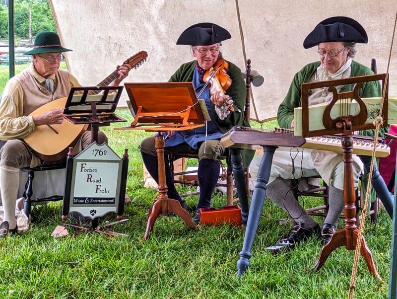 Three musicians dressed in colonial-era attire performing at Fort Loudoun in Franklin County, Pennsylvania. They are seated under a canopy, playing traditional instruments: a lute, a violin, and a hammered dulcimer. A sign in front of them reads '1760, Forbes Road Folke: Music & Entertainment!'.