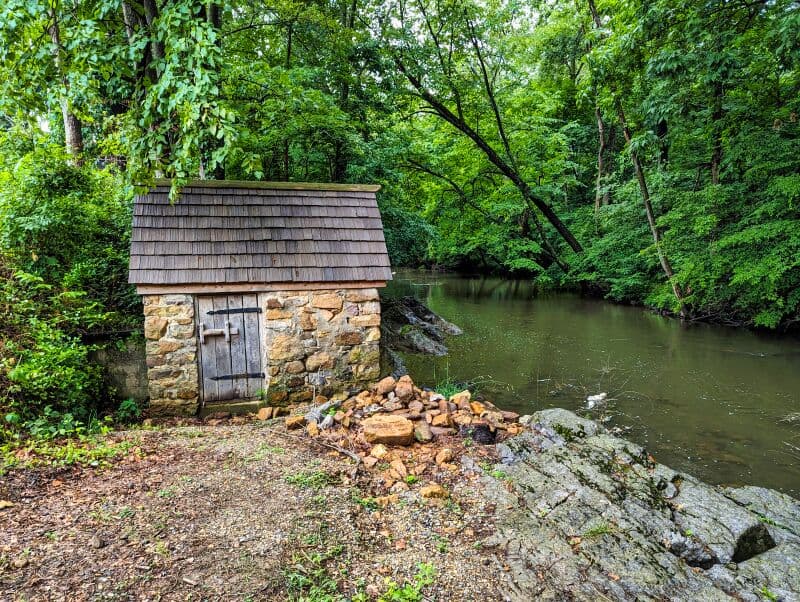Stone-built storage shed with a wooden roof, situated by the edge of a tranquil stream surrounded by dense greenery and trees, at Fort Loudoun in Franklin County, Pennsylvania.