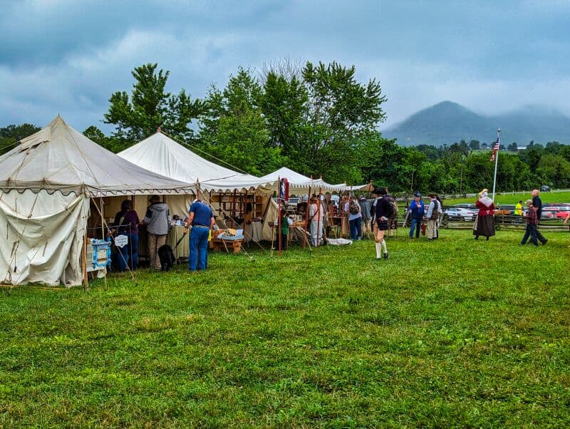 Market Days event at Fort Loudoun in Franklin County, Pennsylvania, featuring people in period costumes interacting amidst white canvas tents set up on a grassy field, with misty mountains and an American flag in the background