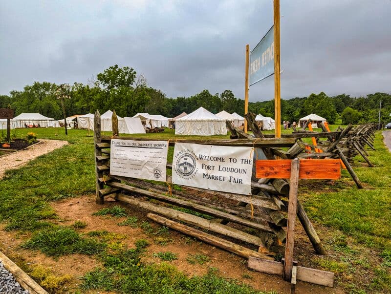 Entrance to the Fort Loudoun Market Fair in Franklin County, Pennsylvania, marked by a wooden barricade displaying banners thanking sponsors and welcoming visitors, with white canvas tents set up in the background on a cloudy day.