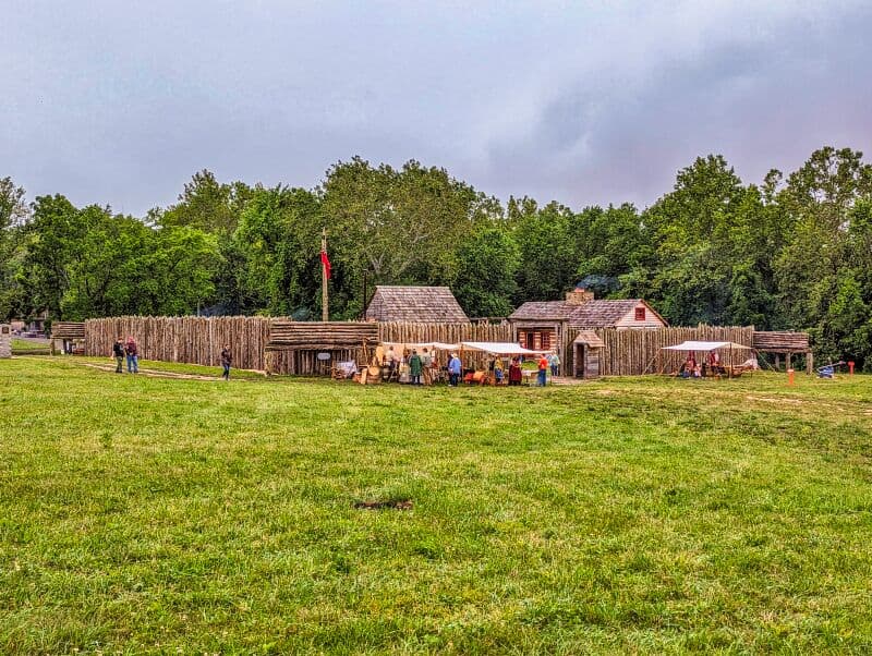 Reconstructed wooden fortifications and buildings of Fort Loudoun in Franklin County, Pennsylvania, with people dressed in period clothing gathering and interacting, set against a backdrop of lush trees on an overcast day.