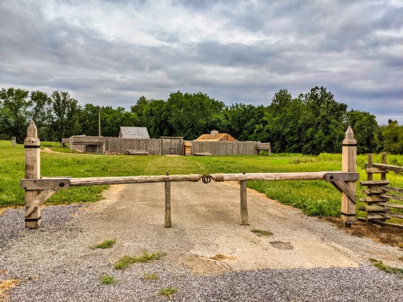 Entrance view of Fort Loudoun in Franklin County, Pennsylvania, with a wooden barrier in the foreground, rustic wooden structures and palisades in the background, set against lush greenery and a cloudy sky.