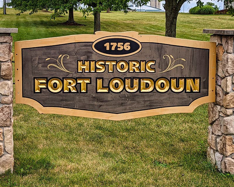 Signage for 'Historic Fort Loudoun' dated 1756, displayed on a wooden plaque with gold lettering, mounted between stone pillars, set against a backdrop of green grass and trees.