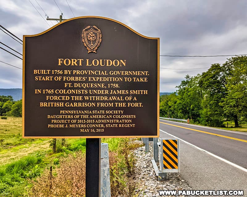Historical marker for 'Fort Loudoun' detailing its construction in 1756 and key events. The plaque is mounted on a post beside a road, with an emblem at the top and information about the Pennsylvania State Society and Daughters of the American Colonists. In the background, there's a road, trees, and a cloudy sky.