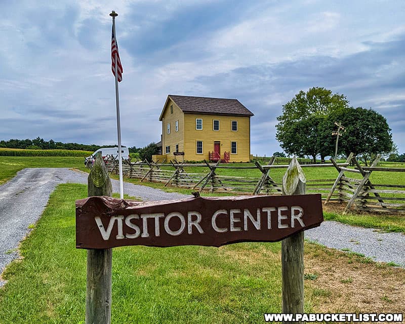 Rustic sign reading 'Visitor Center' on the grounds of Fort Loudoun in Franklin County Pennsylvania, with an American flag flying above it, set against the backdrop of a yellow two-story building, wooden fences, and lush green fields under a cloudy sky.