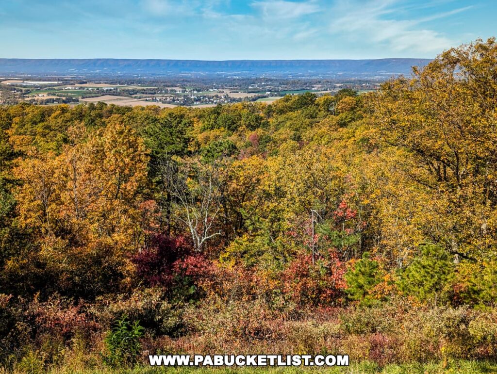 A vibrant autumn forest foreground with a panoramic view of Cumberland Valley and distant Blue Mountain range under a clear blue sky.