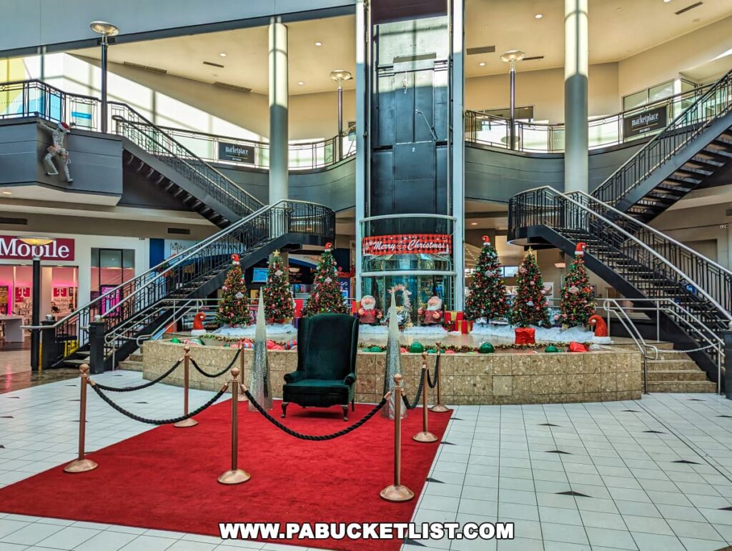 Interior view of the Marketplace an Steamtown in Scranton, Pennsylvania, showcasing a festive Christmas display with multiple decorated trees, presents, and a large Santa chair on a raised platform. The area is cordoned off with a red velvet rope and the floor features a distinctive red carpet. The two-level building has several storefronts and curved staircases on either side leading to the upper level.