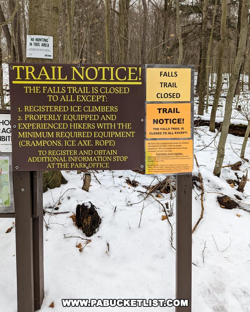 A close-up view of a trail notice sign at Ricketts Glen State Park. The sign states that the Falls Trail is closed to all except registered ice climbers and properly equipped and experienced hikers with the minimum required equipment, which includes crampons, ice axe, and rope. Additional information and registration are available at the park office. A 'No Hunting' sign is also visible in the background, with a snow-covered ground and winter-bare trees surrounding the area.