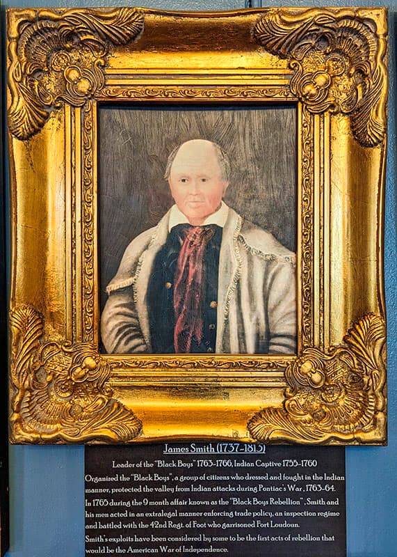 Ornate gold-framed portrait of James Smith (1757-1813) displayed at Fort Loudoun, Franklin County, Pennsylvania. Smith is depicted as an older man with white hair, wearing an 18th-century coat with a fur collar. Below the portrait, an informational plaque describes his significance: 'Leader of the "Black Boys" 1765-1766, Indian Captive 1755-1760. Organized the "Black Boys", defended the valley from attacks during Pontiac's War, and led the "Black Boys Rebellion".' The text further highlights Smith's leadership at Fort Loudoun and his role in early acts of rebellion that led to the American War of Independence.