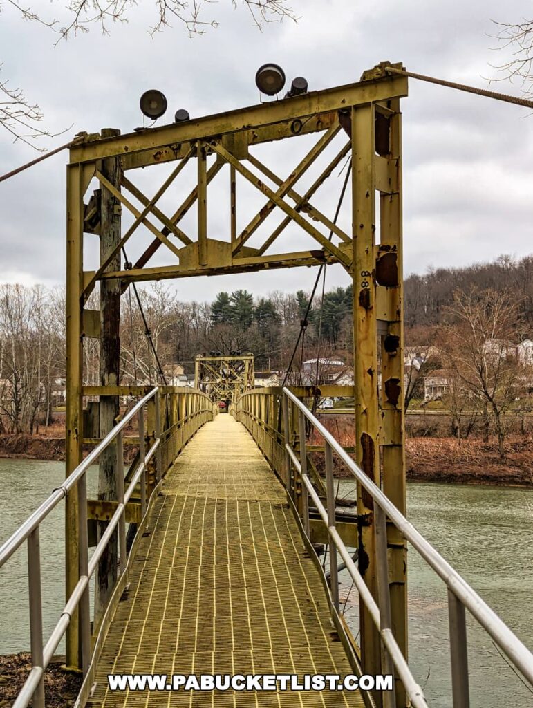 A photograph of the Hyde Park Walking Bridge in Western Pennsylvania, featuring a long, narrow pedestrian suspension bridge with yellow metal trusses and supports, leading across a river. The bridge has a gridded metal walkway and is flanked by metal railings, with trees and a cloudy sky in the background.