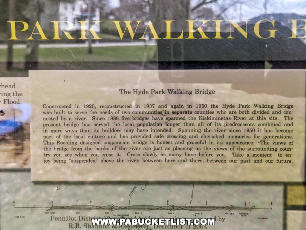 Close-up of an informational plaque about the Hyde Park Walking Bridge in Western Pennsylvania. The text, partially obscured by reflections and a cobweb, details the bridge's history, noting its construction in 1920, reconstruction in 1937 and 1950, and its significance to the local communities. It mentions that the bridge spans the Kiskiminetas River and has been part of local culture and memories for generations.