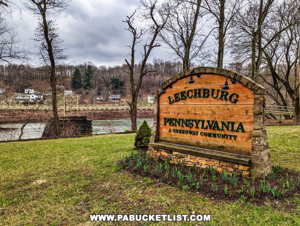 The 'Leechburg Pennsylvania A Greenway Community' sign made of rustic wooden planks with carved and painted lettering, mounted on a stone base and illuminated by overhead lights. Young daffodil plants are emerging from the ground in front, with the Hyde Park Walking Bridge and a river in the background, flanked by leafless trees under a cloudy sky.