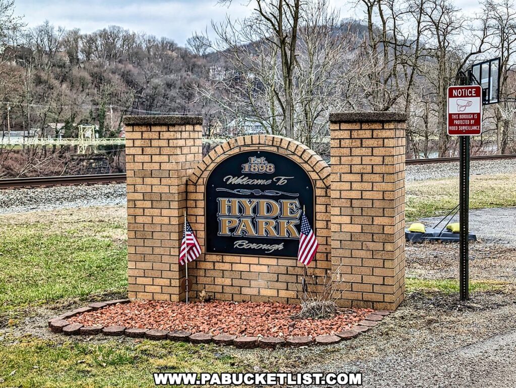 A welcoming sign for Hyde Park Borough, established in 1898, with elegant gold and black lettering on a brick signpost, flanked by American flags. In the background, the Hyde Park Walking Bridge is visible across the river, next to a notice sign stating the borough is under video surveillance, against a backdrop of bare trees and a cloudy sky.