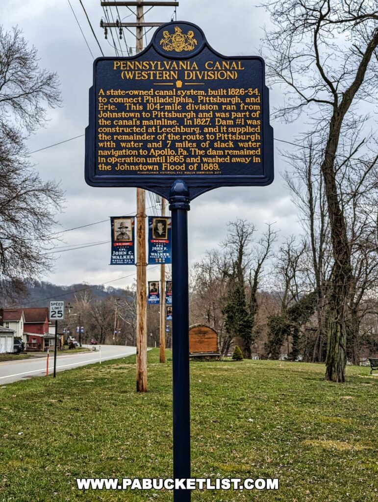 A historical marker sign for the Pennsylvania Canal (Western Division) in Hyde Park, Western Pennsylvania, against a backdrop of leafless trees and an overcast sky. The sign, adorned with the Commonwealth’s coat of arms, provides information on the canal system built between 1826-1834. A wooden covered bridge structure can be seen in the background, alongside a residential street with a speed limit sign.