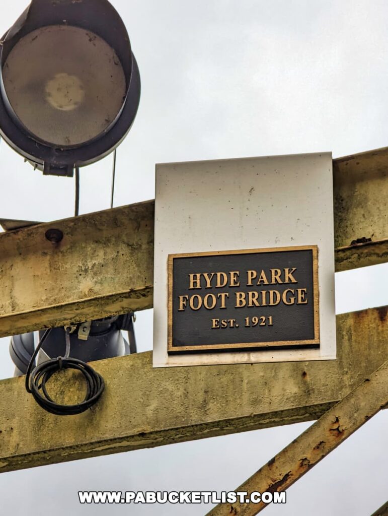 A close-up of a sign on the Hyde Park Foot Bridge in Western Pennsylvania, reading 'HYDE PARK FOOT BRIDGE EST. 1921' in raised, black letters on a weathered metal plaque. Above the sign, there's an old, industrial-style light fixture attached to the yellow-painted metal truss of the bridge, against a grey, overcast sky.