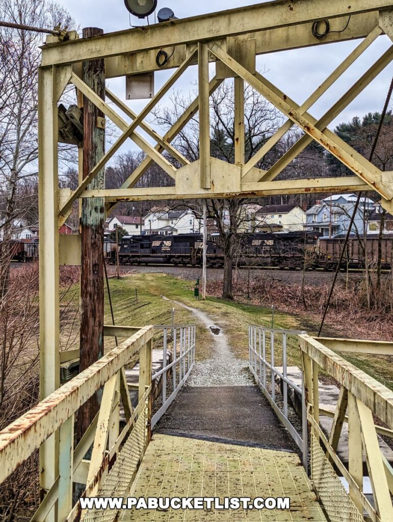 A view from the Hyde Park Walking Bridge in Western Pennsylvania showing a metal truss overhead and the gridded walkway leading towards the far end of the bridge. Beyond the trusses, a freight train with cars labeled 'NS' passes by, juxtaposed against a backdrop of houses and barren trees. The scene captures a moment of stillness on the bridge with the motion of everyday life in the distance.