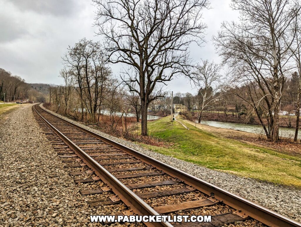 A landscape view of a single railroad track running parallel to a river in Hyde Park, Western Pennsylvania. On the right, the Hyde Park Walking Bridge with its yellow metal structure spans the river, connecting with the lush green riverbank. Leafless trees line both the track and the river, with overcast skies above.