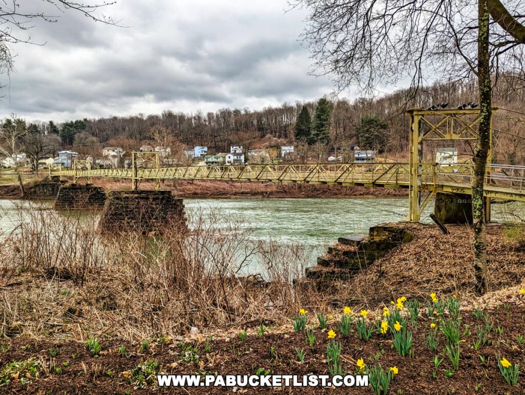 A side view of the historic Hyde Park Walking Bridge in Western Pennsylvania, with yellow daffodils in bloom in the foreground. The bridge, constructed with yellow metal trusses, spans across a river with a backdrop of a residential neighborhood on a hill. Overcast skies and bare trees create a moody early spring atmosphere.