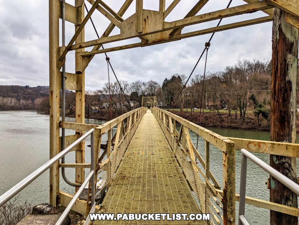 The Hyde Park Walking Bridge in Western Pennsylvania, viewed from the bridge deck itself, showcases the intricate yellow metal truss structure and the suspension cables. The metal grid path of the bridge stretches ahead, flanked by bare winter trees, and overlooks the greenish waters of the Kiskiminetas River on a cloudy day.