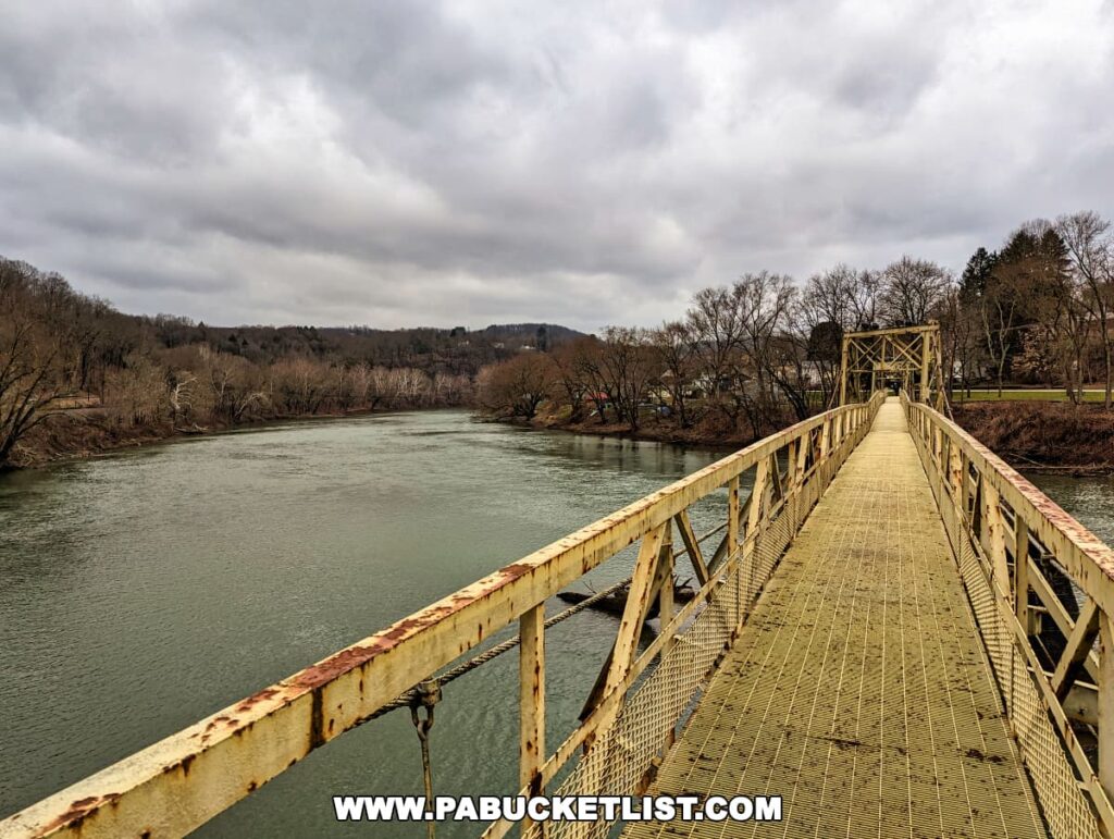 Looking across the Hyde Park Walking Bridge in Western Pennsylvania, the image captures the weathered metal trusses and the mesh walkway that leads across the Kiskiminetas River. The river flows peacefully beneath, surrounded by barren trees and a park in the distance, under a gray, cloud-filled sky.