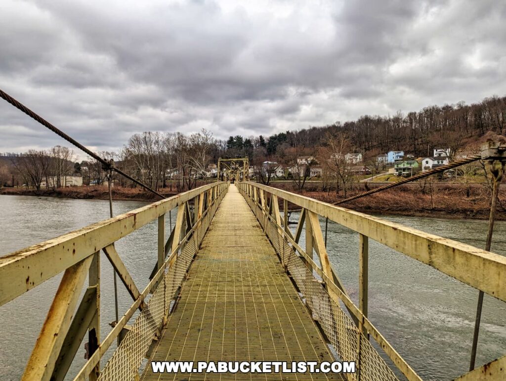 The view from the Hyde Park Walking Bridge in Western Pennsylvania, looking toward Leechburg. The bridge's metal structure frames the walkway, leading the eye toward the small town in the distance. The Kiskiminetas River flows below, flanked by bare trees and houses, under a cloudy sky that hints at the changing seasons