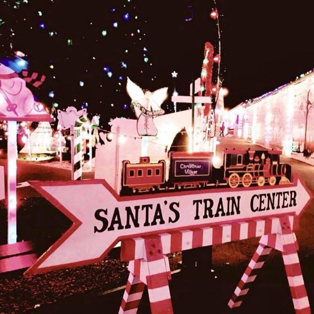 A festive sign at night reading 'Santa's Train Center' with an arrow pointing towards the attraction, situated among colorful Christmas lights and decorations, including a toy train and an angel figure, at Koziar's Christmas Village in Berks County, Pennsylvania.