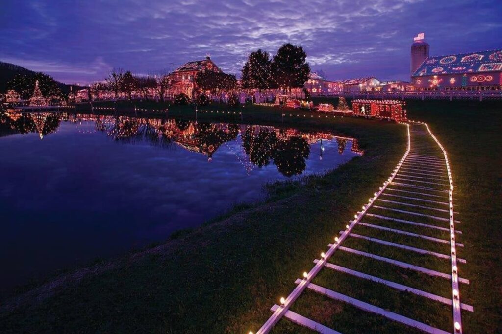 Twilight view of Koziar's Christmas Village in Berks County, Pennsylvania, with a serene pond reflecting the dazzling lights of decorated buildings and trees. A lit path leads the eye through the tranquil scene, enhancing the magical holiday atmosphere.