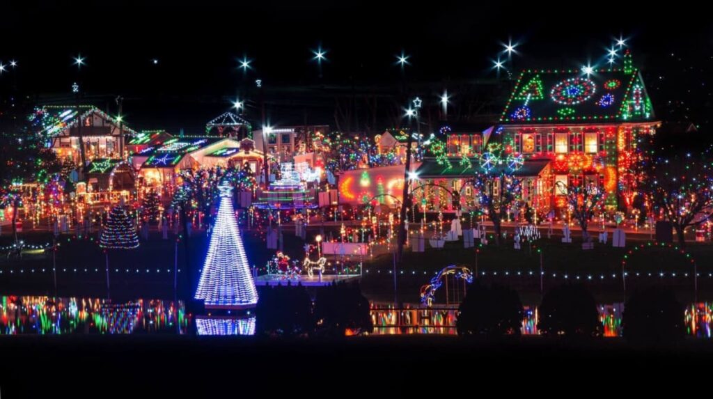A vibrant night scene at Koziar's Christmas Village in Berks County, Pennsylvania, featuring an array of colorful holiday lights reflected in a still water body. The display includes a prominent cone-shaped light structure resembling a Christmas tree, surrounded by illuminated houses and decorations, creating a mirror image on the water's surface.