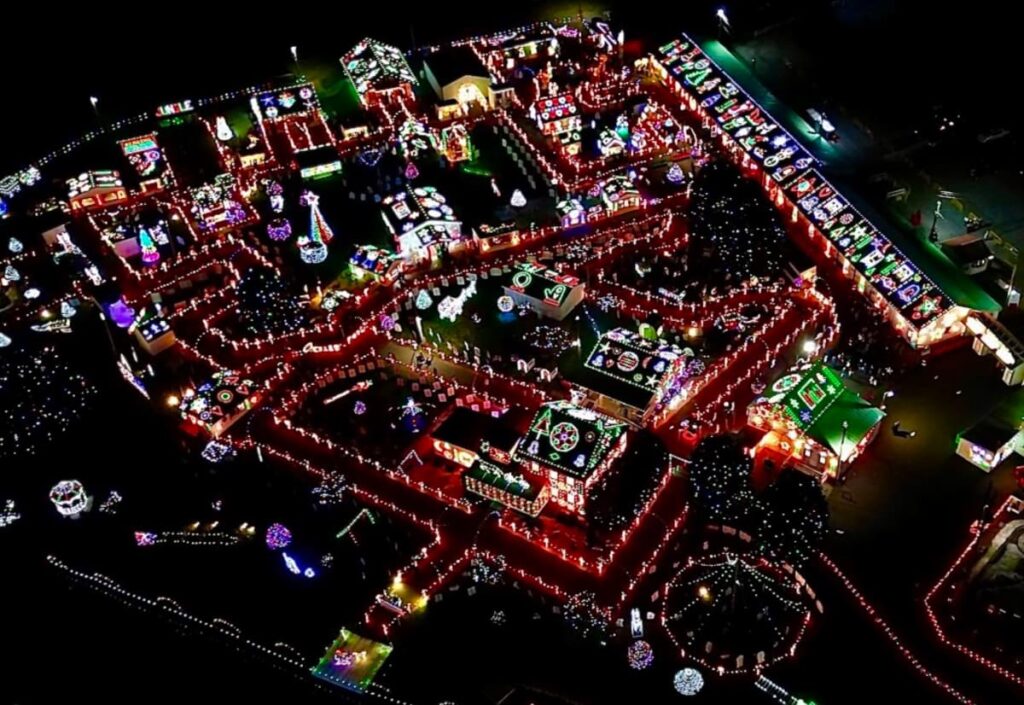Aerial night view of Koziar's Christmas Village in Berks County, Pennsylvania, where the intricate layout of the village is outlined by strings of bright lights. The buildings and paths are adorned with colorful holiday decorations, creating a luminous maze of festive cheer.