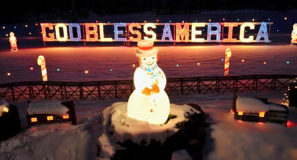 A cheerful snowman figure glows warmly in the foreground at Koziar's Christmas Village in Berks County, Pennsylvania, with a backdrop of large, illuminated letters spelling out 'GOD BLESS AMERICA'. The scene is set against a snowy landscape with a candy cane decoration to the side, creating a patriotic and festive winter scene.