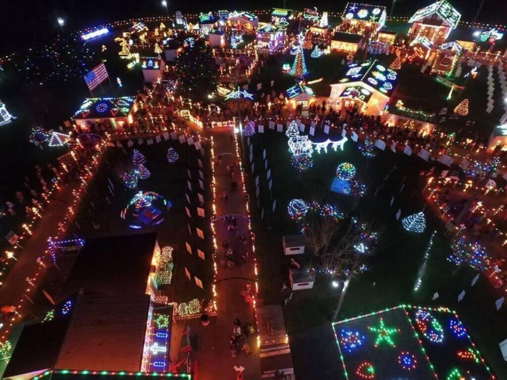 An expansive aerial view at night of Koziar's Christmas Village in Berks County, Pennsylvania, with buildings and pathways intricately outlined in glowing holiday lights. Various festive figures and decorations can be seen throughout, and a large American flag is prominently displayed in lights on a rooftop, adding to the celebratory display.