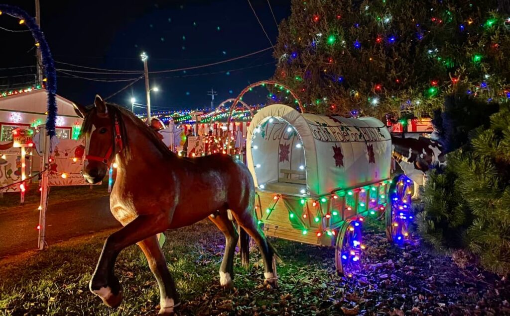 A lifelike statue of a horse stands in the foreground at Koziar's Christmas Village in Berks County, Pennsylvania, beside a festively lit covered wagon with the words 'Toy Ride Drop Off' illuminated. The wagon is surrounded by Christmas lights and evergreen trees, contributing to the enchanting holiday atmosphere of the village.