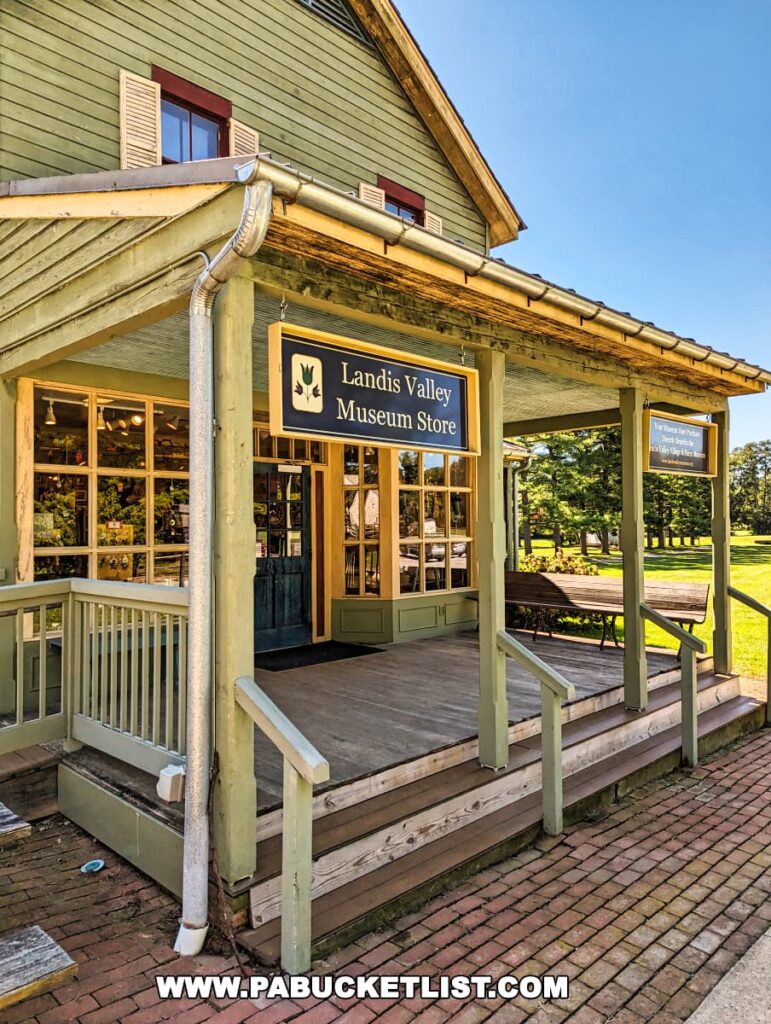 The front porch of the Landis Valley Museum Store in Lancaster, Pennsylvania, showing a pale yellow wooden building with white trim around the windows and the porch. A sign with a logo featuring a bird in flight hangs above the porch, which has a gray floor and is enclosed by a railing.