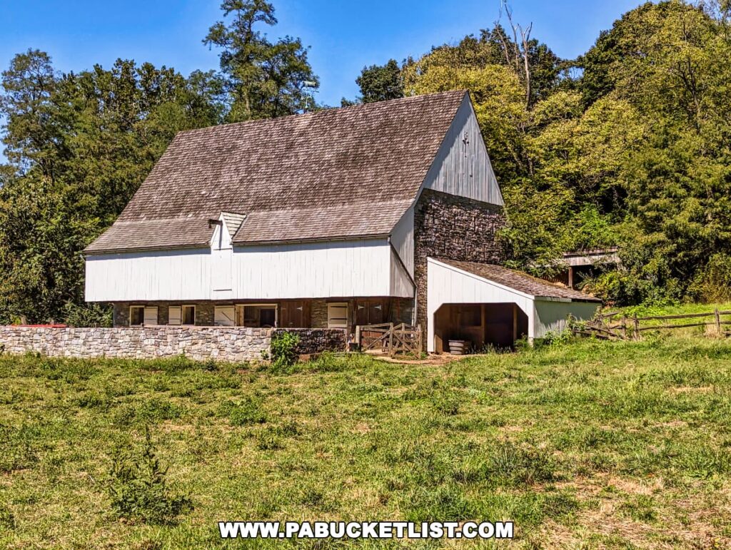 A traditional white Pennsylvania bank barn with a stone foundation at the Landis Valley Museum in Lancaster County, Pennsylvania. The barn is set against a backdrop of lush green trees and a clear blue sky.