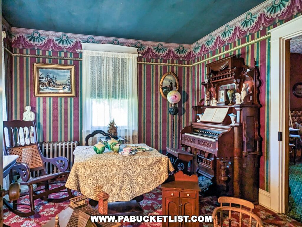 A richly decorated parlor room at Landis Valley Museum in Lancaster, Pennsylvania, showcasing a late 19th-century Victorian interior. The room features dark green and burgundy striped wallpaper with matching fabric valances and drapes. Antique furniture includes a rocking chair, a round table with a lace tablecloth, and an ornate pump organ against the wall. Vintage decorations and a framed landscape painting enhance the historic ambiance.