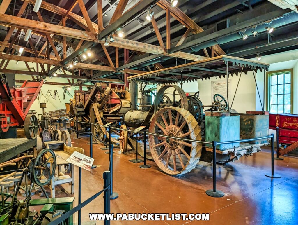 An exhibit within the Landis Valley Museum in Lancaster, Pennsylvania, displaying a collection of antique agricultural machinery, including a large steam tractor with metal wheels. The spacious room has exposed wooden beams and is well-lit, highlighting the historical farming equipment of various sizes and functions, some of which are painted in red.