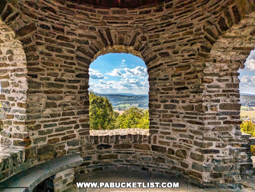 View from inside a circular stone structure at the Marie Antoinette Scenic Overlook in Bradford County, Pennsylvania, with a wide arched opening framing a vast landscape of green fields, trees, and a river in the distance under a blue sky with scattered clouds.