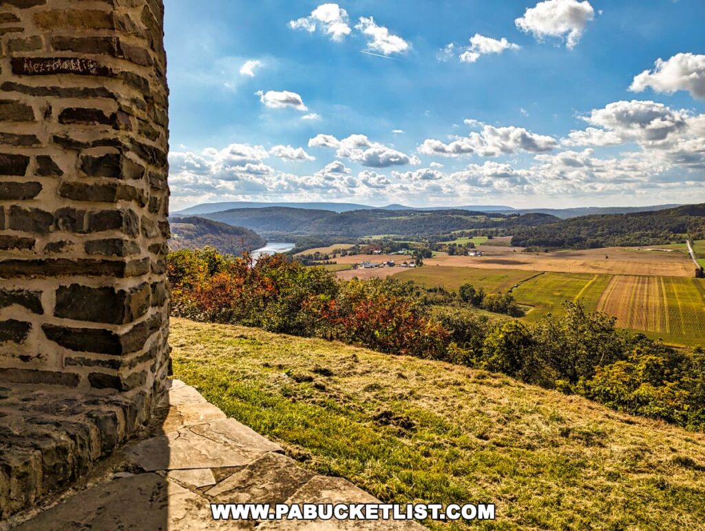 A serene vista from the Marie Antoinette Scenic Overlook in Bradford County, Pennsylvania, framed by the rustic stone edge of the lookout point. The view opens up to a picturesque valley with a river winding through, flanked by rolling hills and scattered farmlands under a partly cloudy sky.