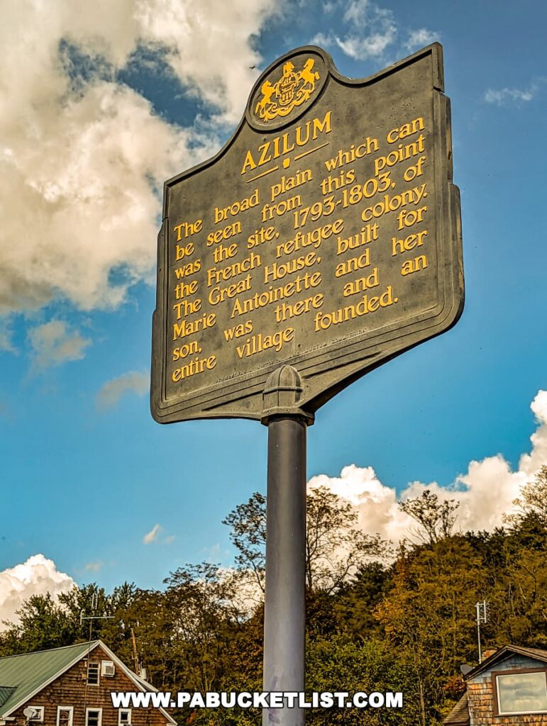 Historical marker titled 'Azilum' under a blue sky with clouds, located at the Marie Antoinette Scenic Overlook in Bradford County, Pennsylvania. The sign, with golden embossed lettering, explains the significance of the location as the site of a 1793-1803 French refugee colony, where a great house was built for Marie Antoinette and her son.