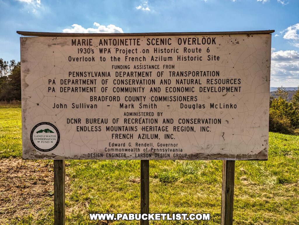 Informational sign at the Marie Antoinette Scenic Overlook in Bradford County, Pennsylvania, detailing the overlook's history as a 1930s WPA project on Historic Route 6 and its relation to the French Azilum historic site. The sign lists various Pennsylvania state departments, county commissioners, and conservation entities involved in funding and administration, against a backdrop of a blue sky with fluffy clouds and a green landscape.