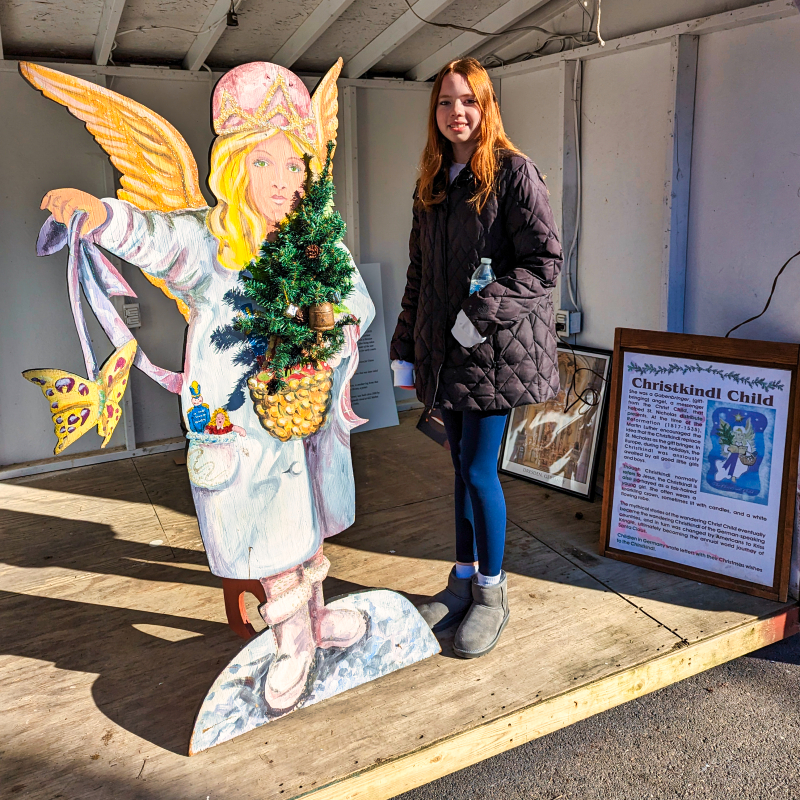 A visitor standing beside a colorful life-size cutout of the Christkindl angel, holding a Christmas tree and a butterfly, with an informational poster about the Christkindl Child in the background, at the Mifflinburg Christkindl Market in Pennsylvania.